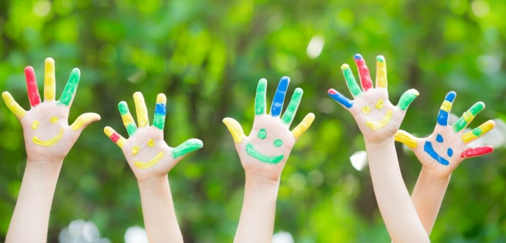 Kinderhände mit bunten Smileys und bunt bemalten Fingern vor grünem Blätterhintergrund
